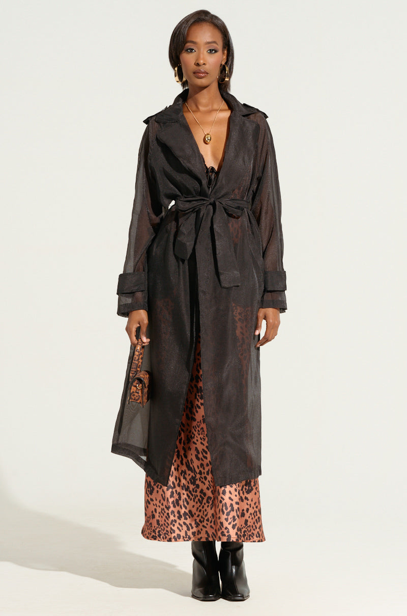 A woman stands facing the camera, wearing the CHIFFON CHATEAU LONG TRENCH IN BLACK tied at the waist over a leopard print dress. She accessorizes with black ankle boots, gold earrings, and a necklace. Her hair is straight and she holds a small leopard print bag, embodying an autumnal layering style with a femme fatale finish.