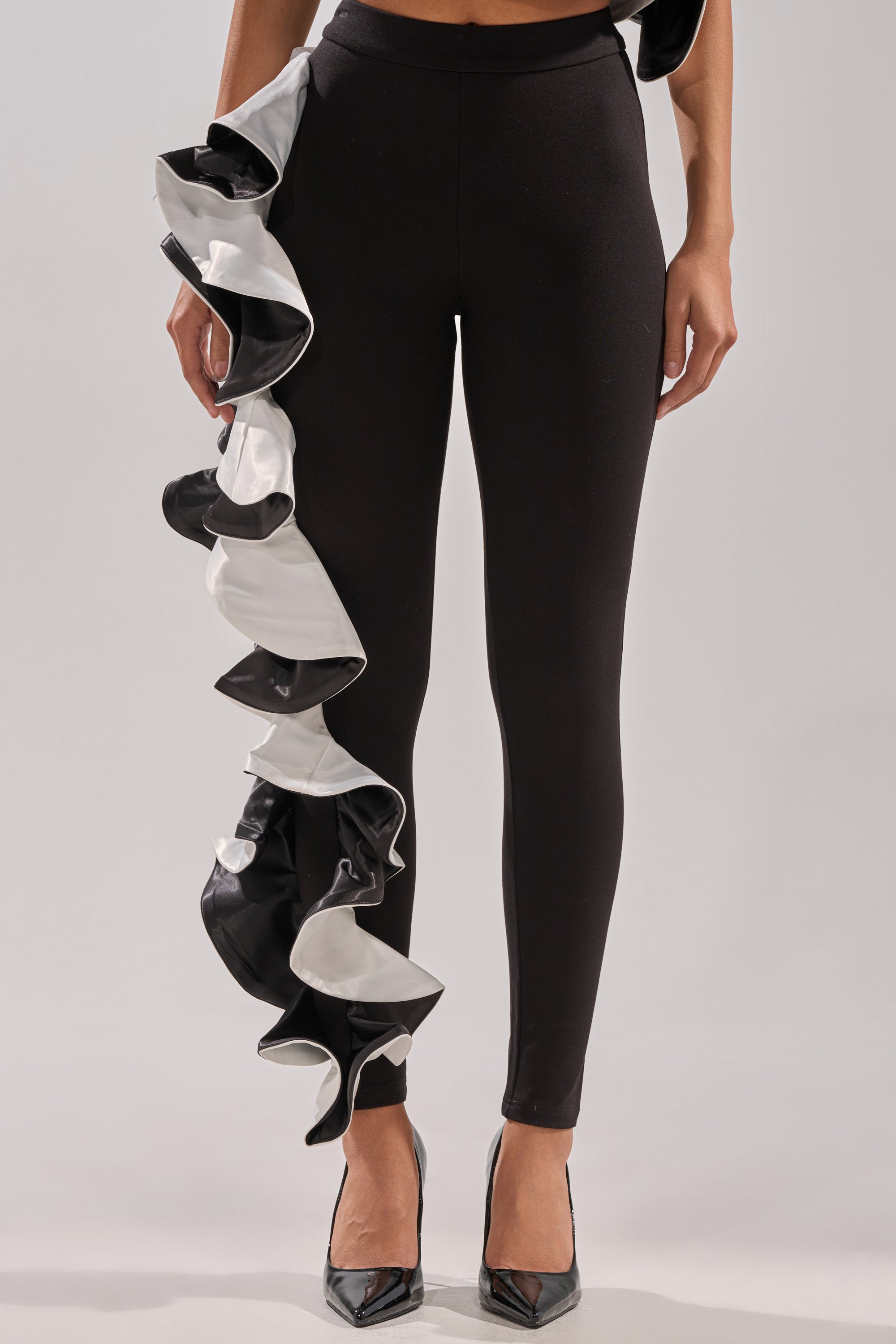 A person wearing CLASSY AND FAB FASHION PANTS, featuring a bold black design with a cascading ruffle down one leg, paired with pointed black heels.