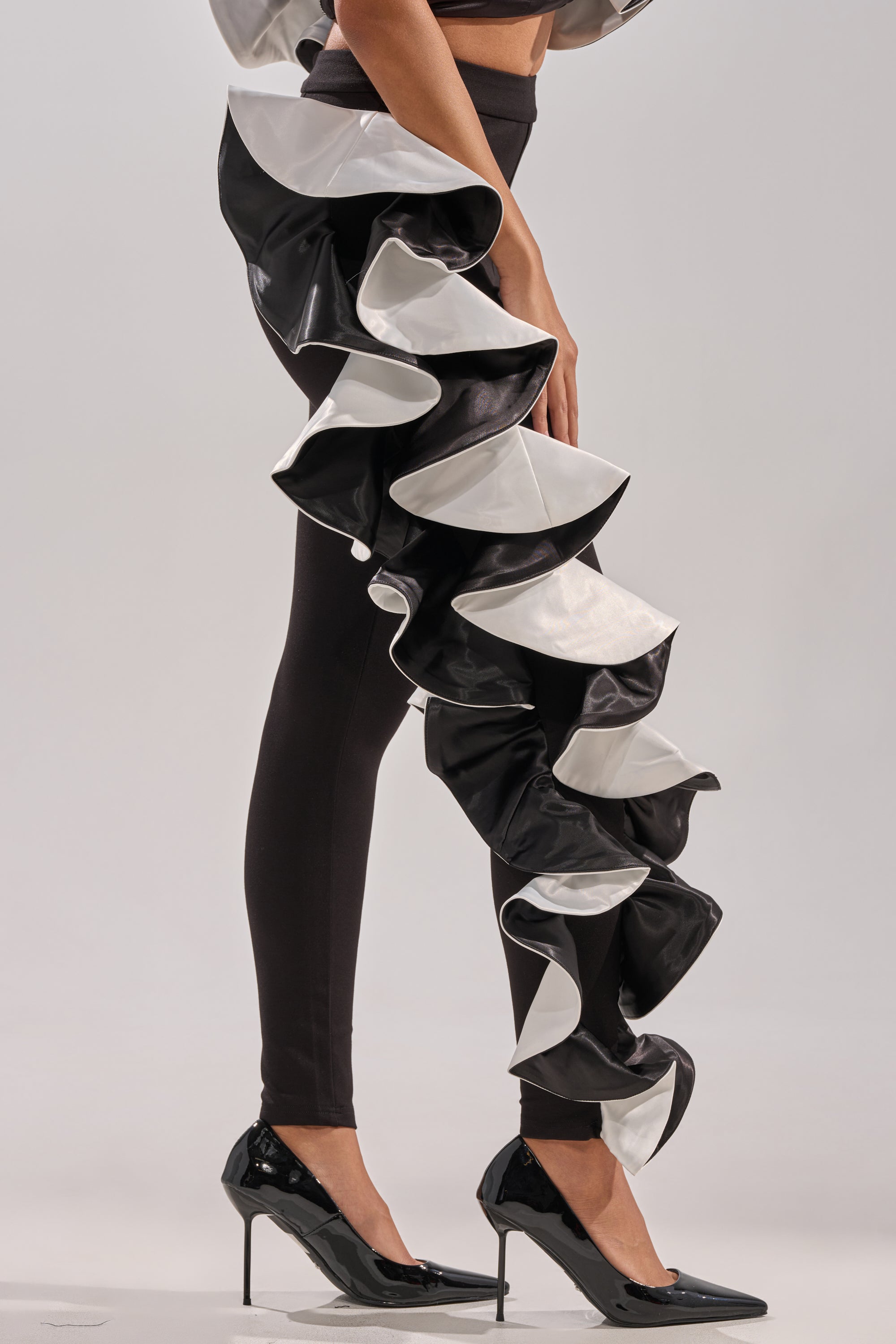 A person wears CLASSY AND FAB FASHION PANTS featuring dramatic black-and-white ruffles down one leg, paired with black high heels, making a bold fashion statement against a plain, light-colored background.