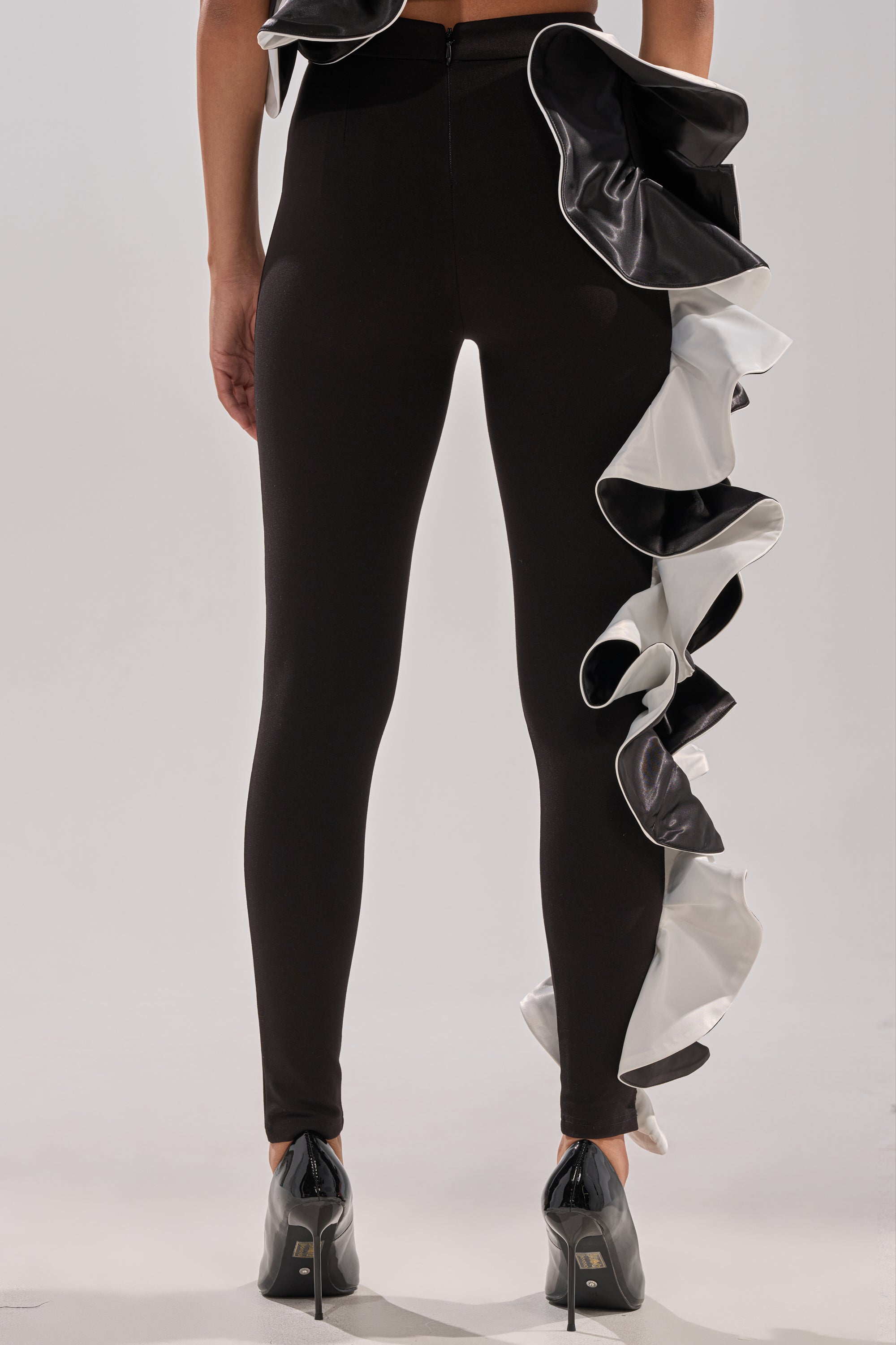 A person faces away in CLASSY AND FAB FASHION PANTS—black fitted pants featuring an asymmetrical black and white ruffle trim—paired with black heels. The plain, light background highlights this high-fashion statement piece.