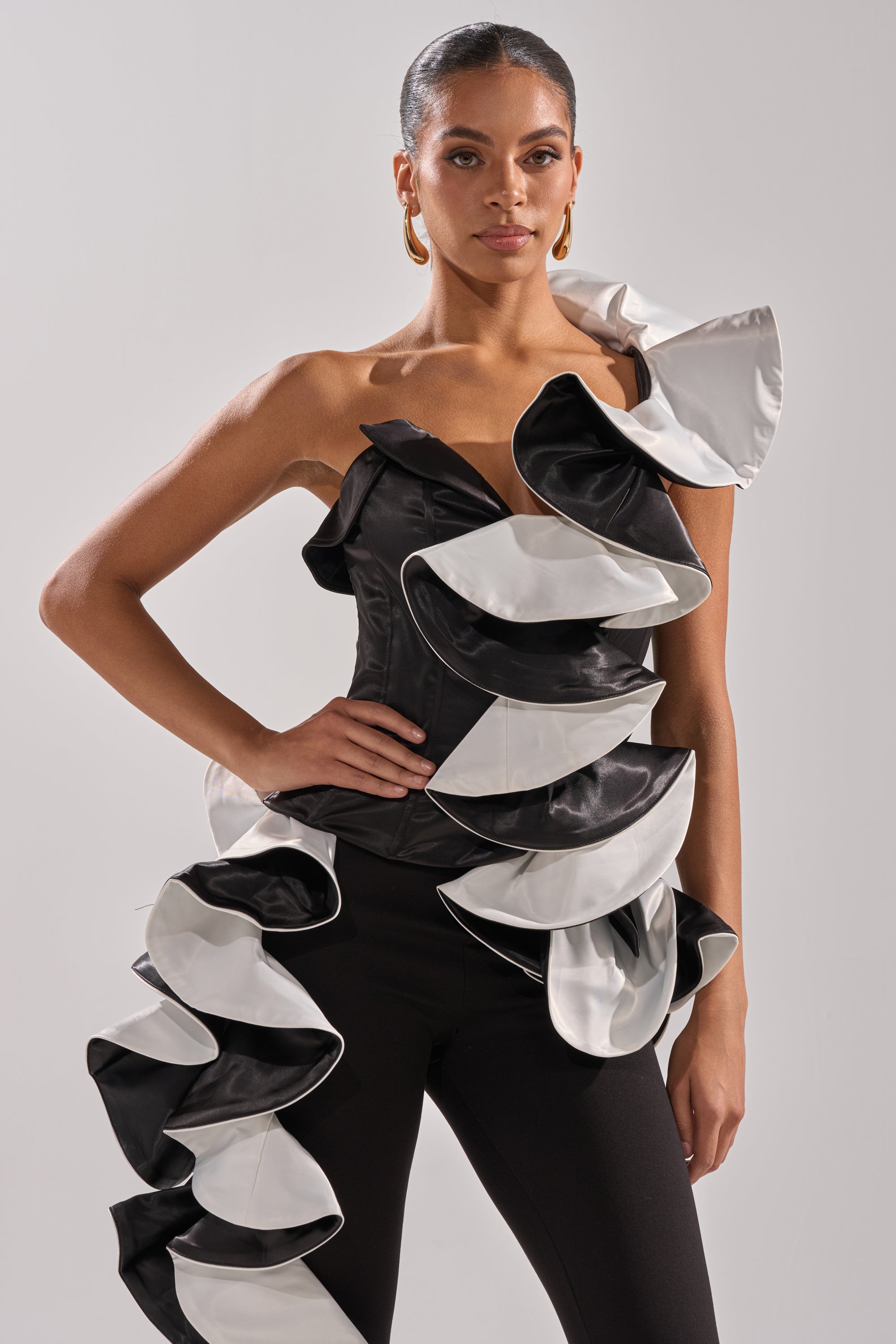 A woman stands confidently against a plain light background, wearing the SNOB IS A HABIT RUFFLE DETAIL SATIN TOP with sculptural ruffles, paired with black pants. Her hair is pulled back and gold hoop earrings complete her chic look.