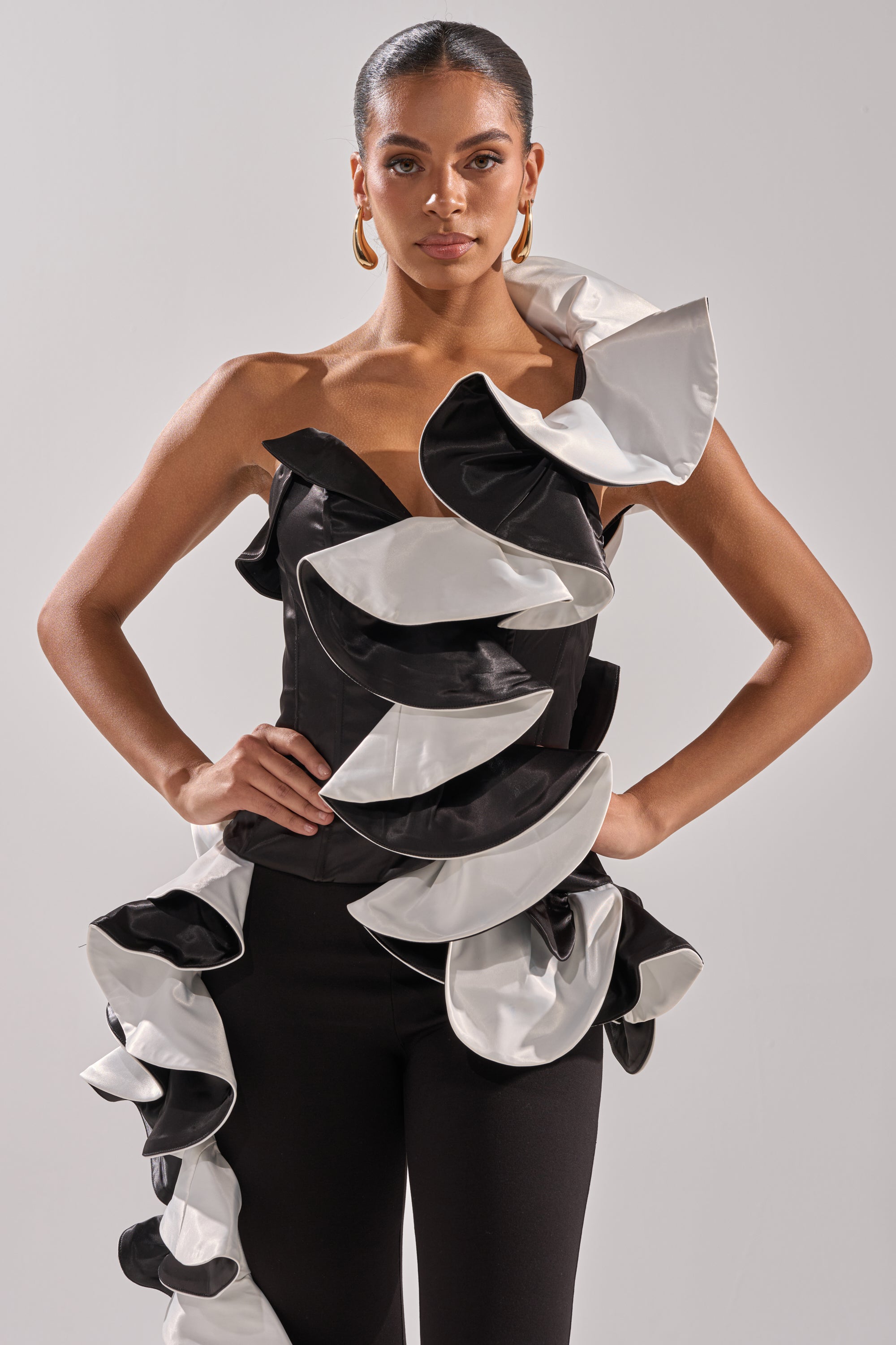 A woman poses confidently in the SNOB IS A HABIT RUFFLE DETAIL SATIN TOP, a couture-inspired black satin one-shoulder piece with dramatic ruffles, paired with gold hoop earrings and slicked-back hair against a light gray background.