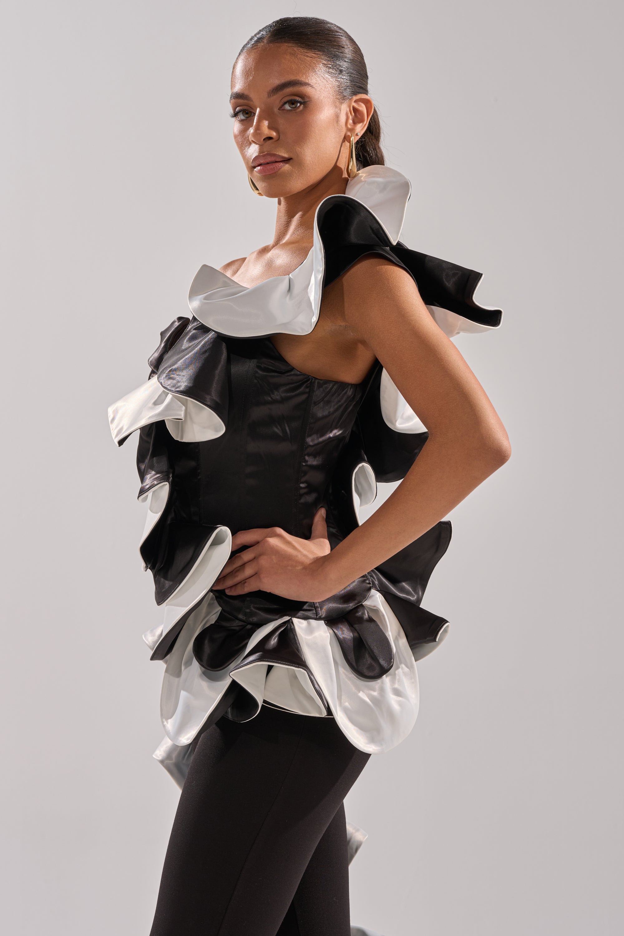 A woman poses confidently against a plain light background, wearing the SNOB IS A HABIT RUFFLE DETAIL SATIN TOP with couture-inspired black and white sculptural ruffles, paired with hoop earrings and dark pants.