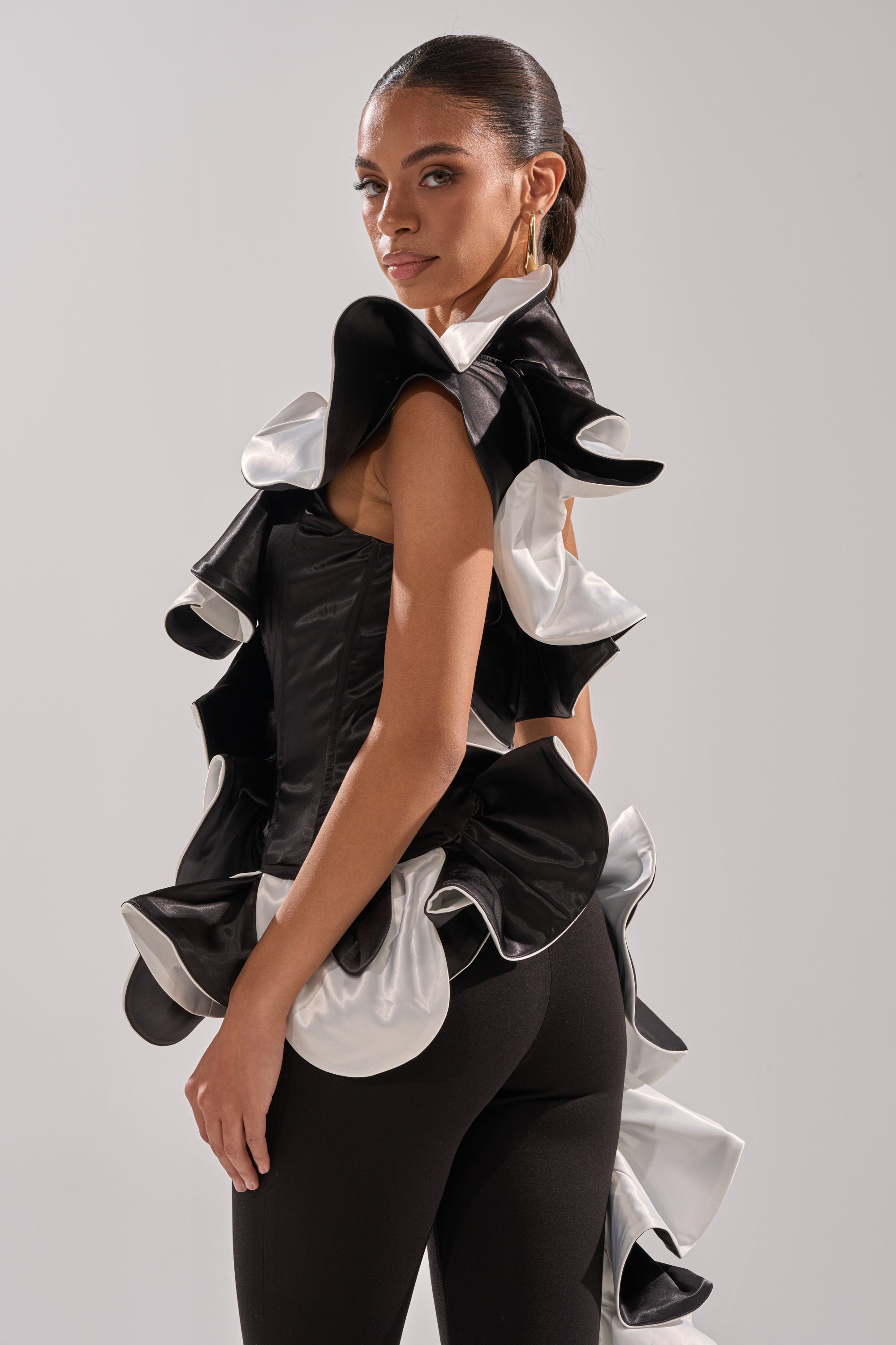A woman models the SNOB IS A HABIT RUFFLE DETAIL SATIN TOP, featuring couture-inspired ruffled edges, paired with black fitted pants. She poses against a plain light gray background with slicked-back hair and hoop earrings.