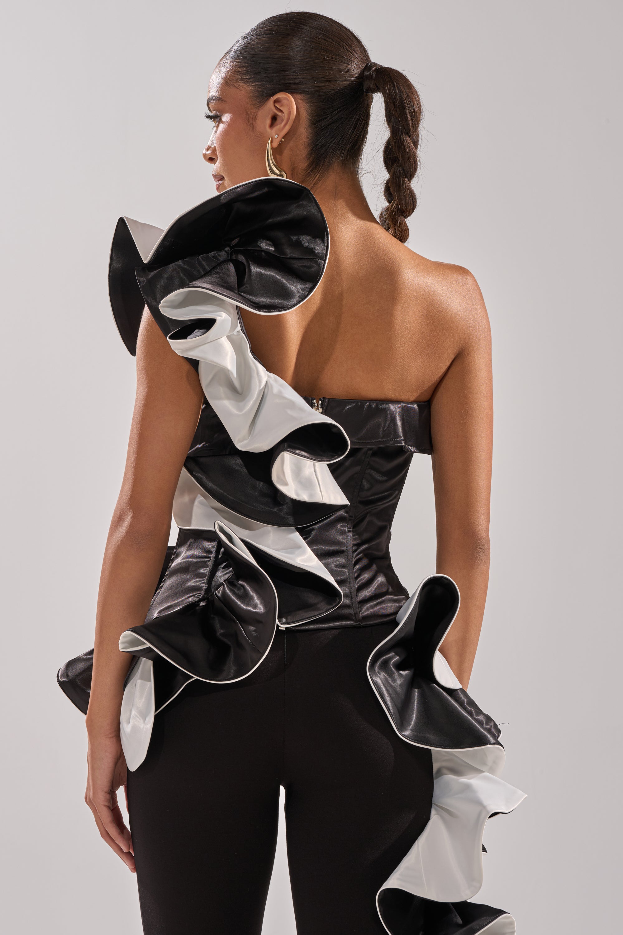 A woman wears the SNOB IS A HABIT RUFFLE DETAIL SATIN TOP, a couture-inspired black satin blouse with dramatic ruffled black and white accents on the side and shoulder, paired with fitted black pants and a sleek, braided ponytail.