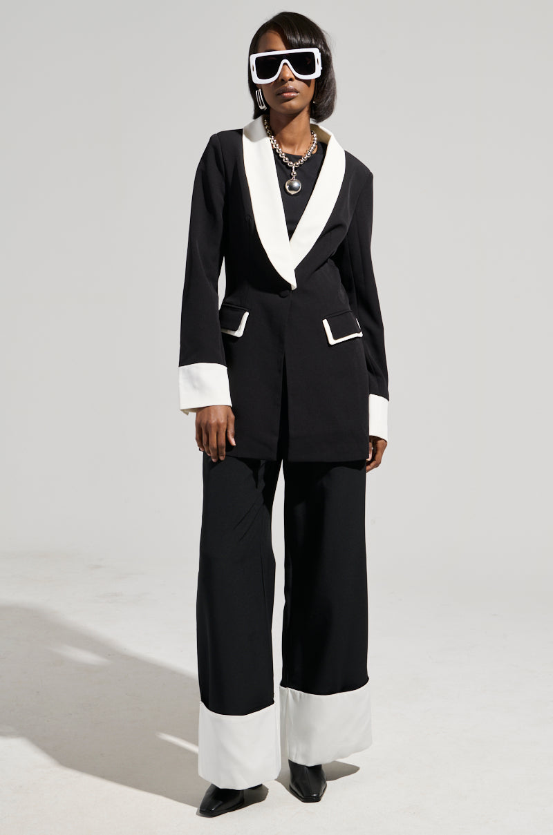 A person wearing a sleek, stylish suit with the MIDNIGHT IN PARIS TROUSER—a black and white outfit featuring wide-legged pants—poses against a plain background. The non-stretch fabrication of the suit is complemented by white lapels and pocket accents. To elevate the look, they accessorize with large, white-framed sunglasses and a layered necklace.