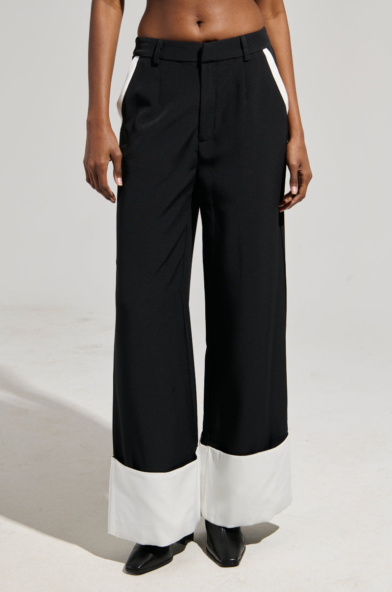 The person is wearing the MIDNIGHT IN PARIS TROUSER, featuring a black, high-rise waist design with a wide-leg silhouette and white cuffs that highlight its non-stretch fabric. Complemented by sharp black pointed shoes, this ensemble pops against the neutral backdrop, emphasizing its chic details.