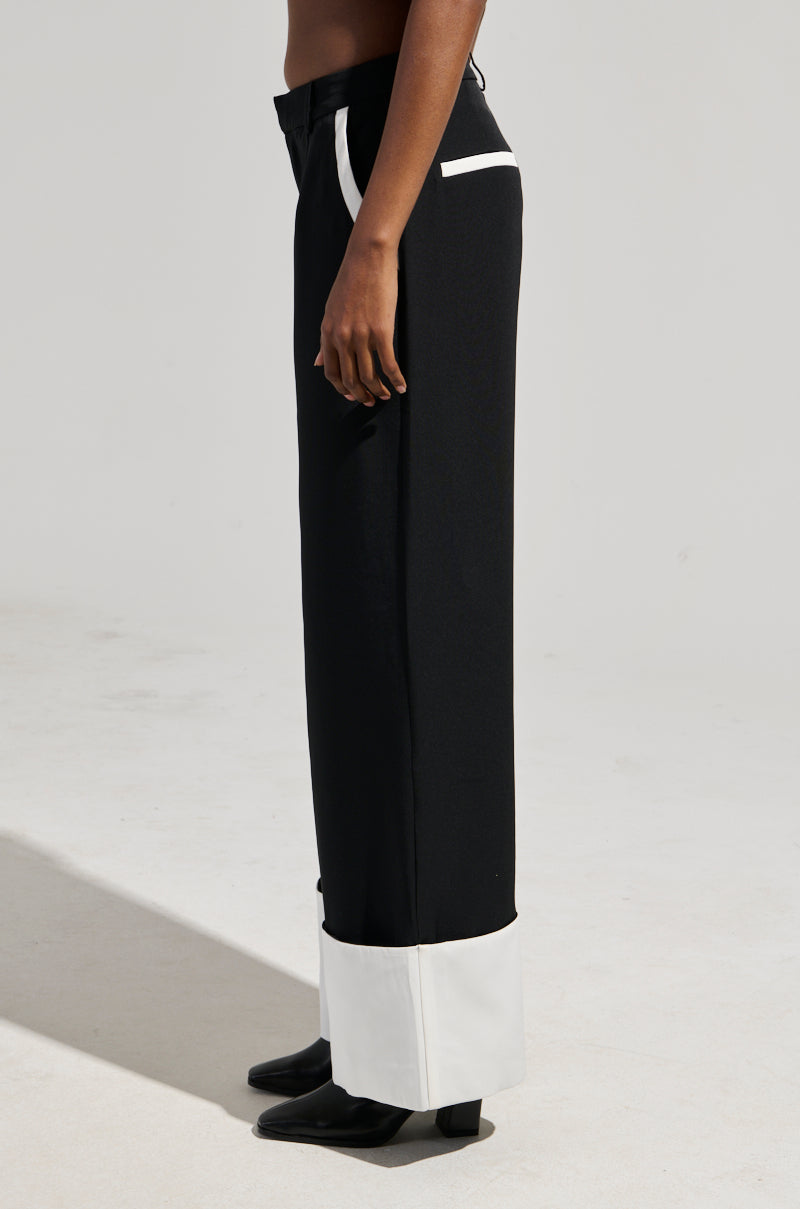 A person wearing the MIDNIGHT IN PARIS TROUSER, featuring black fabric with white cuffs and side pocket details, showcases a sleek high-rise waist. The non-stretch material beautifully enhances the look, elegantly paired with black shoes. A plain, light-colored background highlights the trouser's timeless appeal.