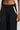 A person is depicted from the waist down against a light gray background, wearing the MIDNIGHT IN PARIS TROUSER. These black, wide-leg trousers feature white stripes on the sides and a high-rise waist. The non-stretch fabric provides structure as their right hand hangs relaxed at their side.