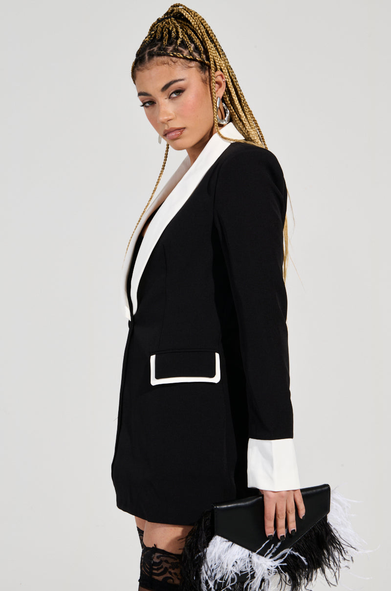 A person with braided hair wears the chic Midnight In Paris Contrast Trim Fitted Blazer in black, displaying bold white accents. They carry a black clutch embellished with delicate white feather details and accessorize with hoop earrings, set against a plain background.