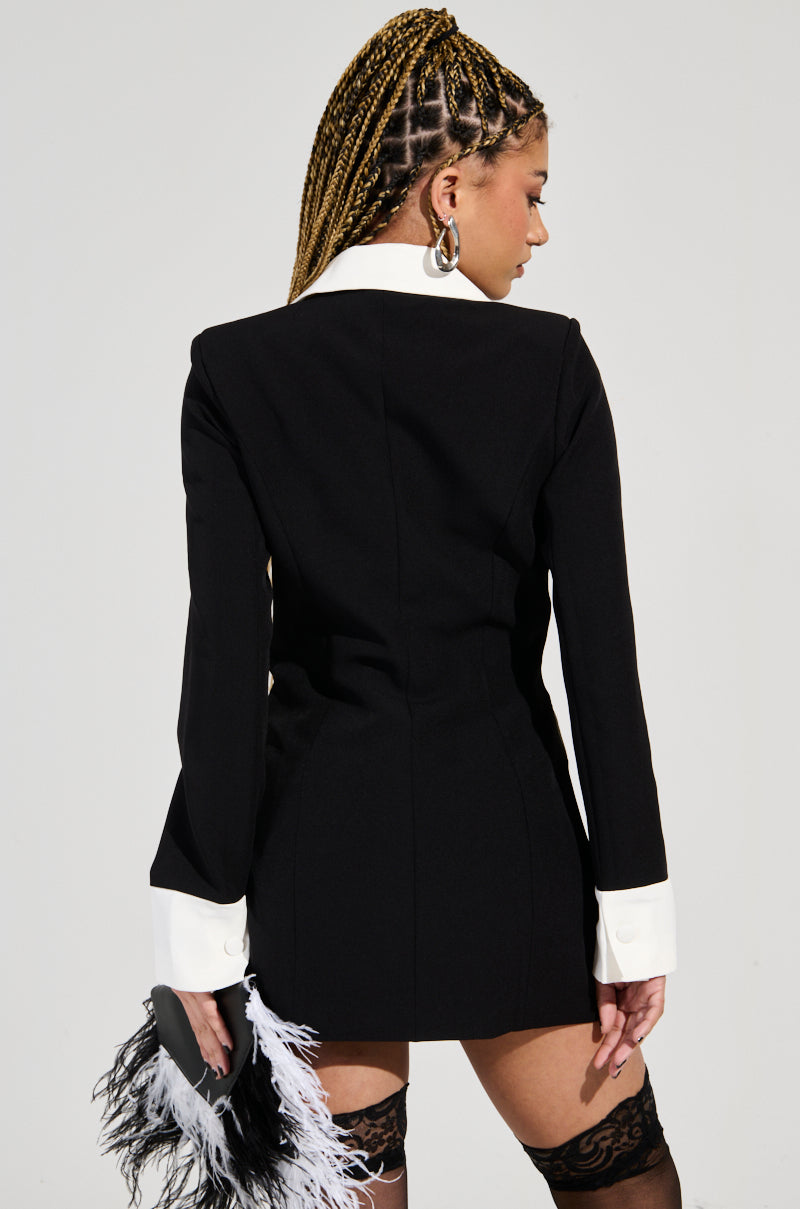 A person with braided hair is wearing a chic Midnight In Paris Contrast Trim Fitted Blazer in Black, holding a small black clutch adorned with white feathers. The individual is turned away, showcasing the back of the outfit, and has paired it perfectly with lace stockings for an elegant touch.
