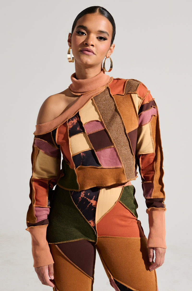 A woman stands against a plain background wearing the CATCH FEELINGS PATCHWORK SWEATSHIRT, a stylish mock neck, off-shoulder outfit in earthy shades of orange, brown, pink, and green. Crafted from stretchy cotton fabrication, she sports gold hoop earrings and her dark hair is tied back. She looks confidently at the camera.