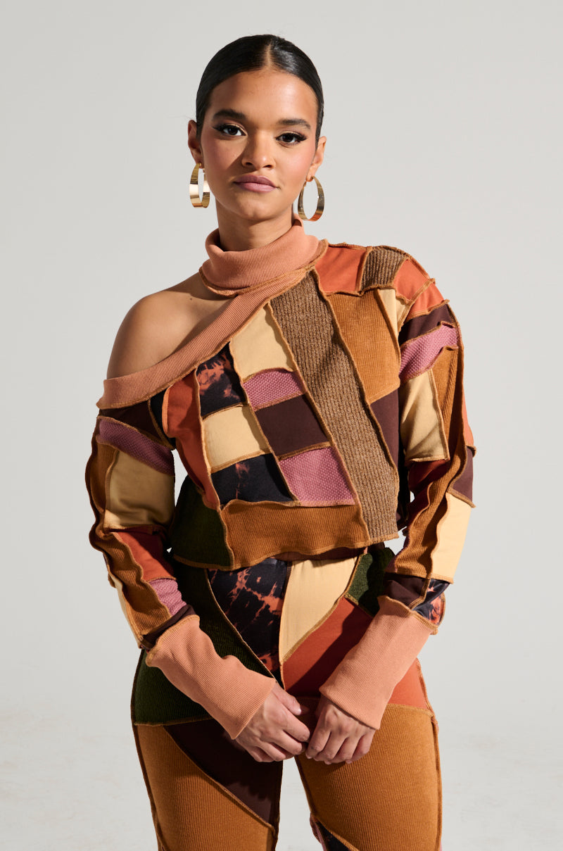 A person stands confidently, wearing a stylish CATCH FEELINGS PATCHWORK SWEATSHIRT from the AKIRA Label, showcasing a mix of textures. The top features an asymmetrical design with one shoulder exposed. They sport large hoop earrings and have their hair neatly pulled back, with a plain and neutral background complementing the look.