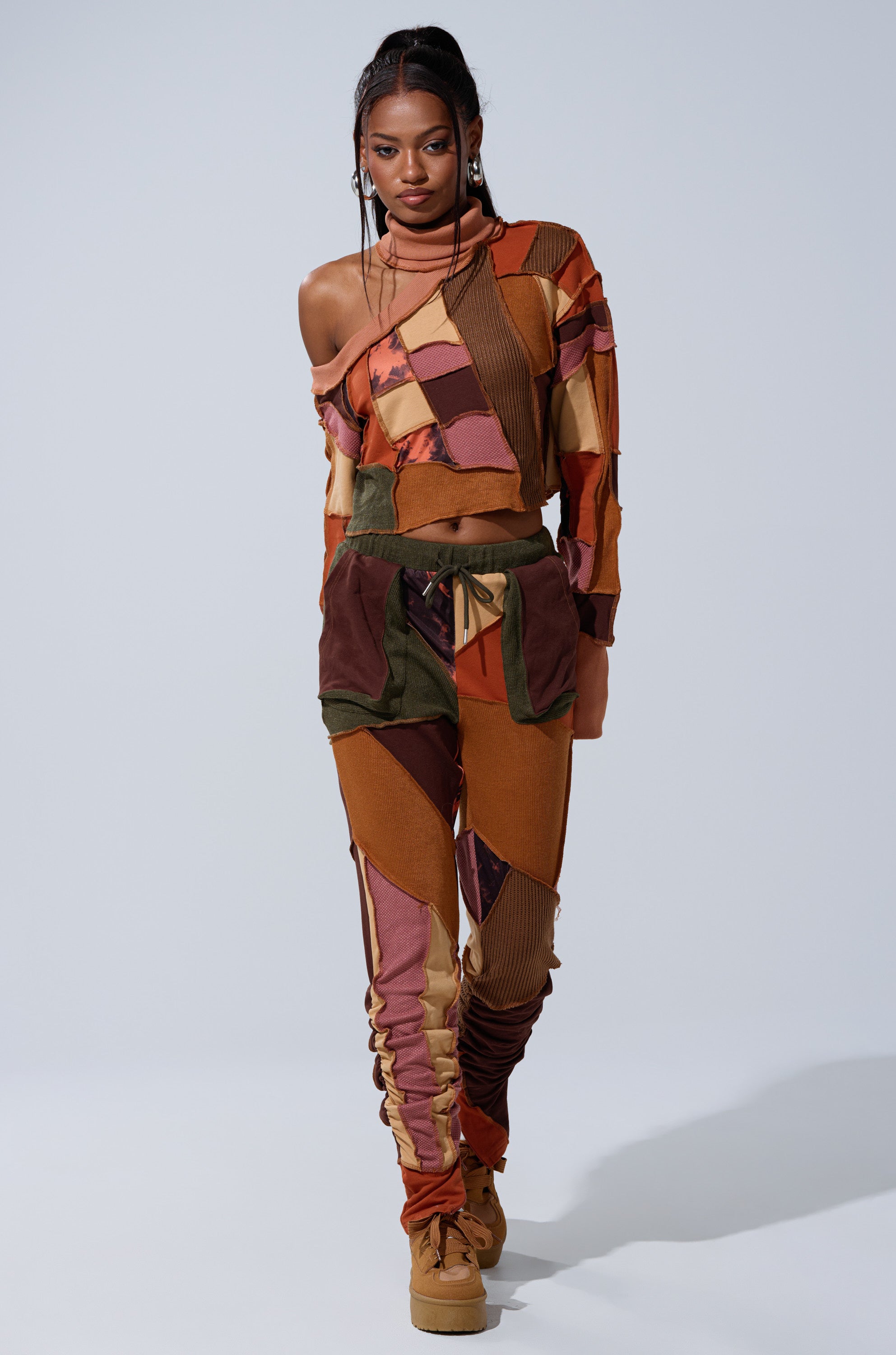 A woman poses against a plain backdrop, wearing earth-tone patchwork leggings called the CATCH FEELINGS PATCHWORK LEGGING, styled with a matching off-shoulder top and tan boots.