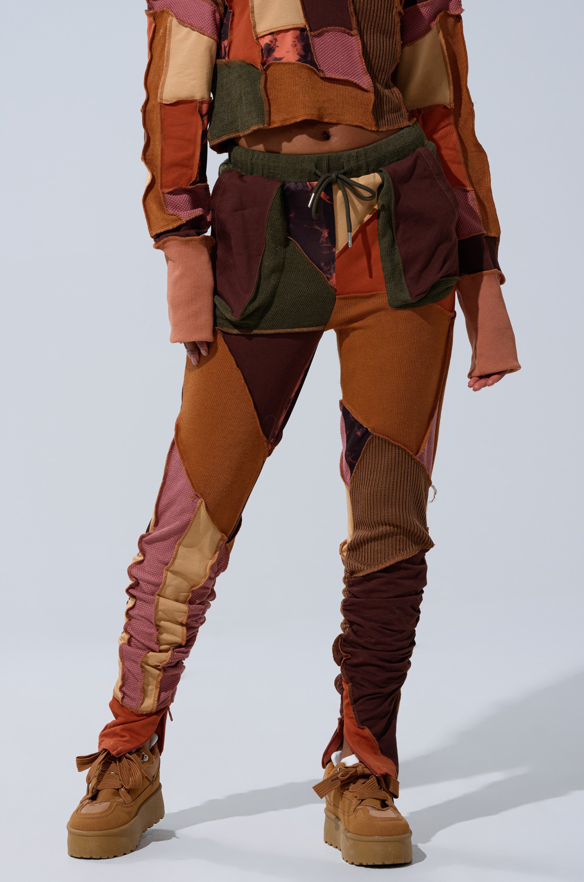 A person wearing the CATCH FEELINGS PATCHWORK LEGGING in a jogger style with a matching cropped top in brown, orange, and red patchwork shades, paired with brown platform shoes.