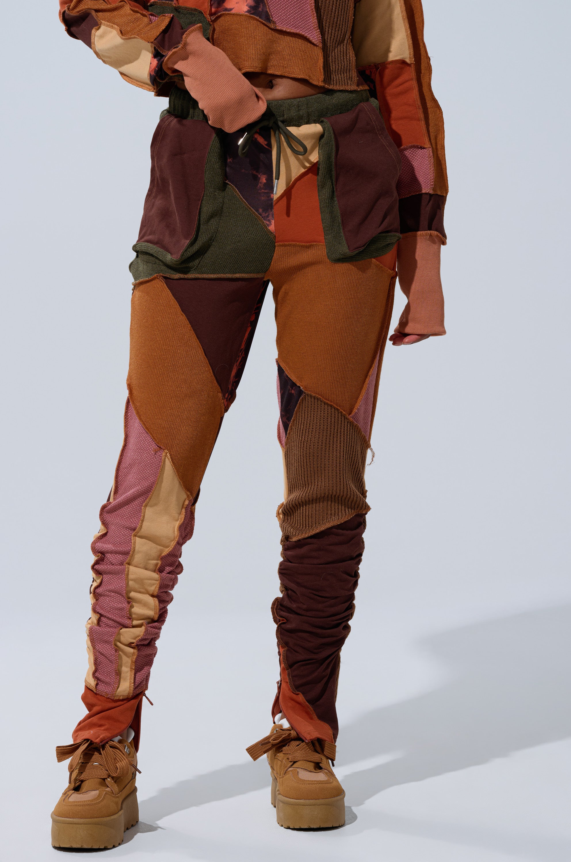 Model wears CATCH FEELINGS PATCHWORK LEGGING and matching top in brown, orange, and maroon with ruched pant legs. Styled with tan platform shoes against a plain, light background.