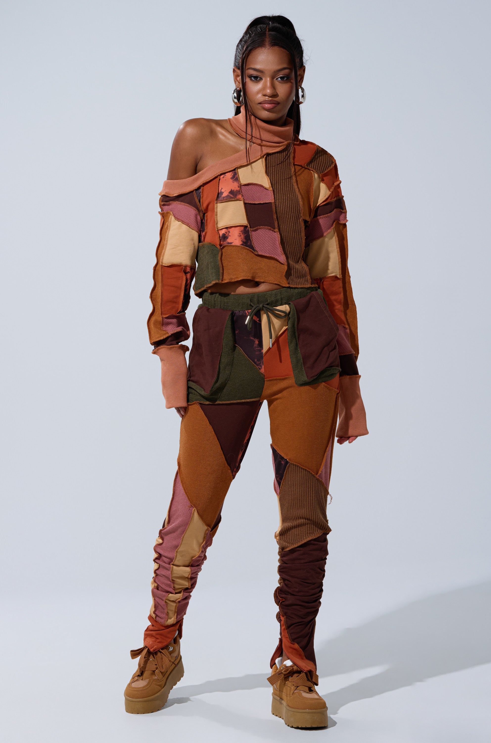 A woman stands confidently in the CATCH FEELINGS PATCHWORK LEGGING, styled with an earthy off-shoulder top and tan platform shoes, set against a plain light background.