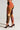 Person wearing colorful CATCH FEELINGS PATCHWORK JOGGER pants from the AKIRA Label and dark brown high-heeled shoes, standing sideways on a plain background. The pants feature various fabrics in earthy tones such as orange, brown, and green.