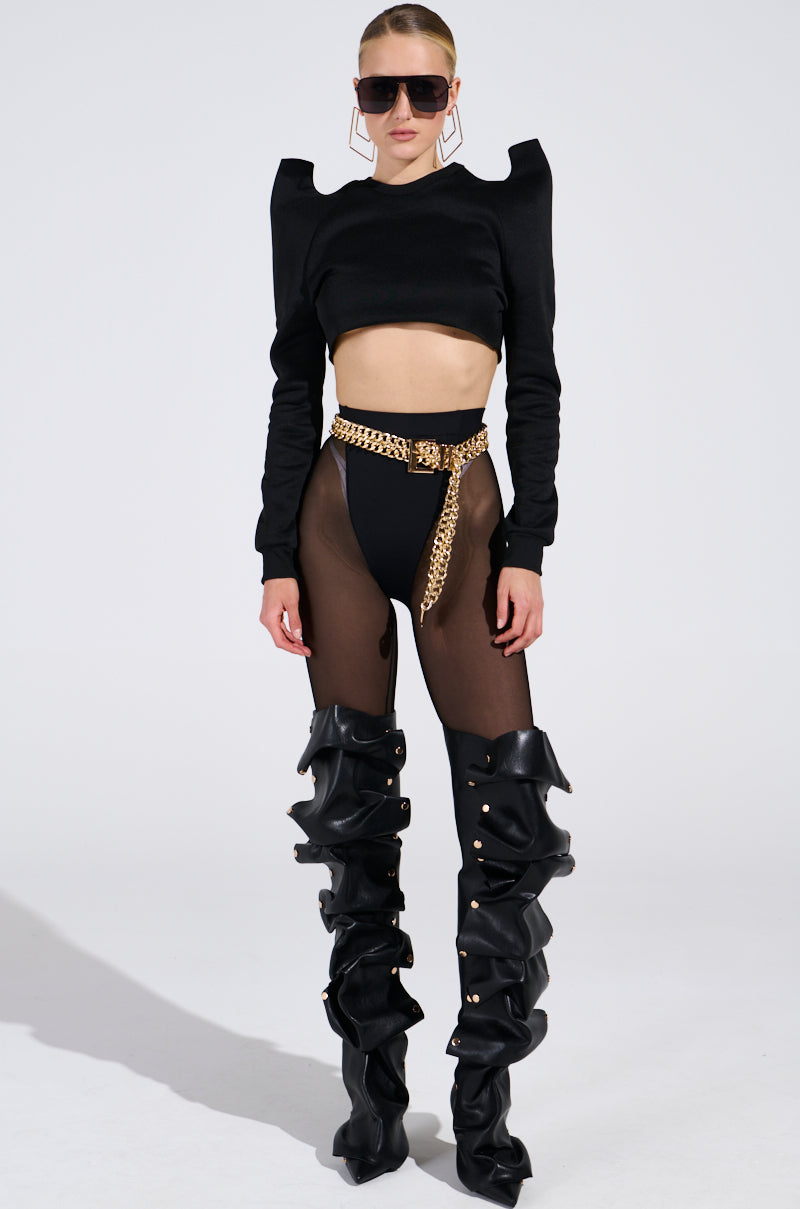 A woman stands confidently in a chic ensemble, wearing a black long-sleeve crop top with structured shoulders, high-rise QUERENDO TE MESH LEGGING with a stretchy waistband, a gold chain belt, and black slouchy thigh-high boots. Her hair is tied back and she sports large sunglasses.