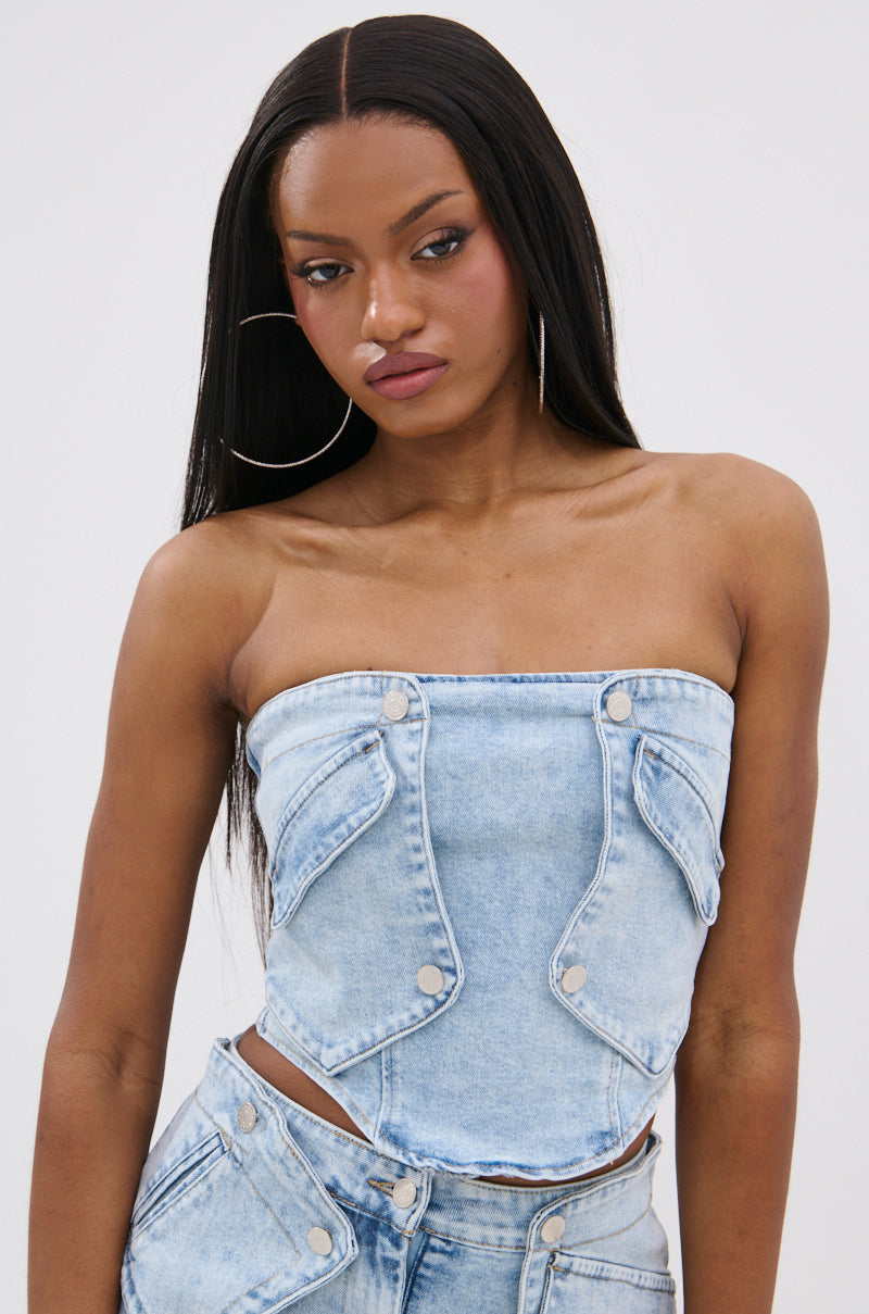 A woman with long straight hair models the OLIVIA DENIM CORSET, a light-wash denim top with pocket details, paired with matching high-waisted bottoms and large hoop earrings. She poses neutrally against a plain white background. 0
