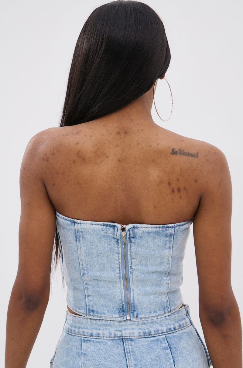 A woman with long straight hair, large hoop earrings, and a tattoo on her upper back wears the OLIVIA DENIM CORSET with a back zipper, matching high-waisted jeans, and has visible acne scars. 3
