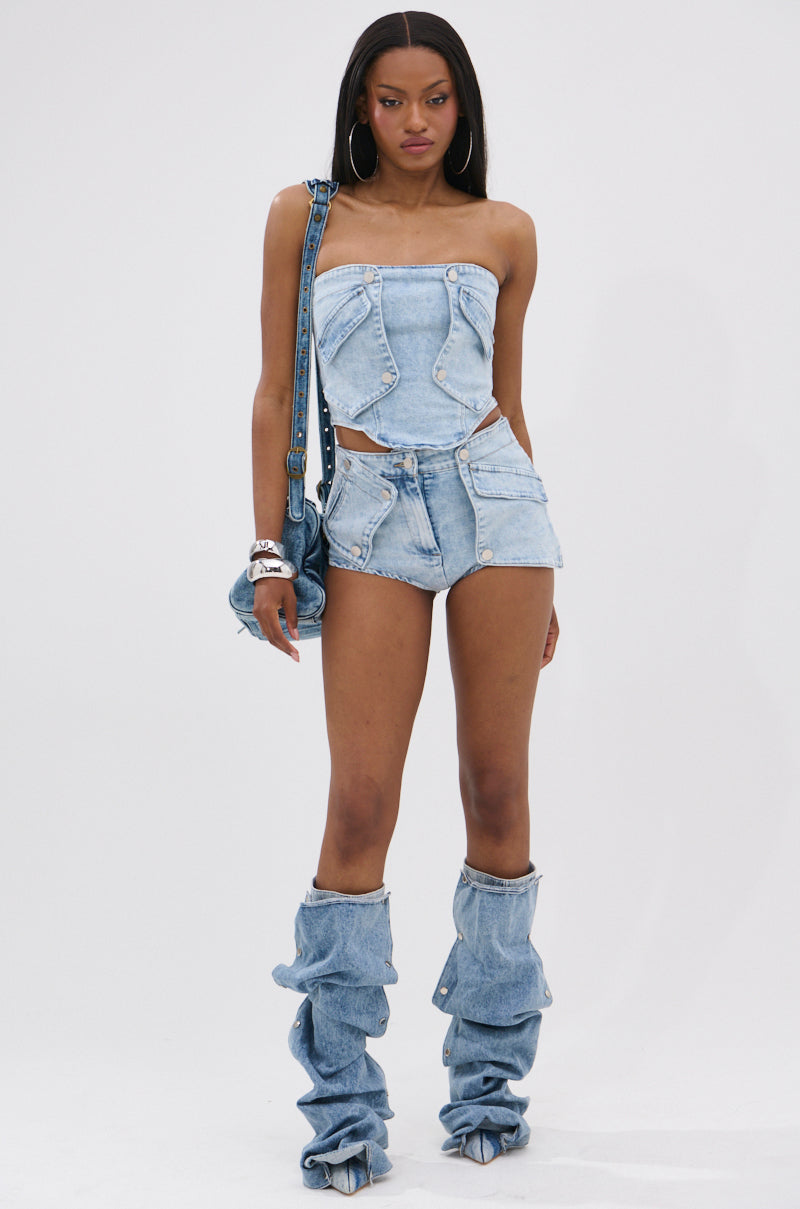 Wearing the OLIVIA DENIM CORSET with matching high-waisted cutout shorts, slouchy denim leg warmers, platform heels, hoop earrings, bracelets, and a denim handbag, a woman poses against a plain white background. 4