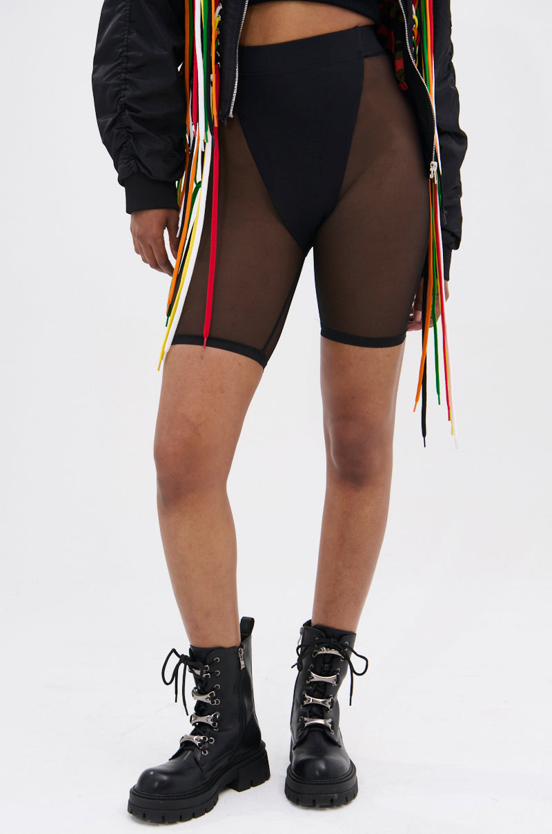A person wears the BARELY THERE MESH BIKER SHORT in black with a high-rise elastic waist, paired with black underwear, lace-up combat boots, and a black jacket featuring colorful ribbon details against a light background.