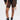 A person wears the BARELY THERE MESH BIKER SHORT in black with a high-rise elastic waist, paired with black underwear, lace-up combat boots, and a black jacket featuring colorful ribbon details against a light background.