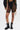 A person wears the BARELY THERE MESH BIKER SHORT in black with a high-rise elastic waist, paired with black underwear, lace-up combat boots, and a black jacket featuring colorful ribbon details against a light background.
