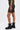 A person models the BARELY THERE MESH BIKER SHORT—black mesh biker shorts with a high-rise elastic waist—paired with a black cropped top and lace-up boots against a plain white background.