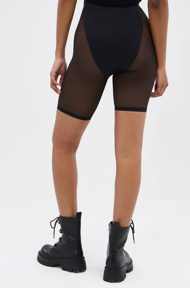 A person stands facing away in BARELY THERE MESH BIKER SHORTS with a high-rise elastic waist, paired with black lace-up combat boots, against a plain light background.