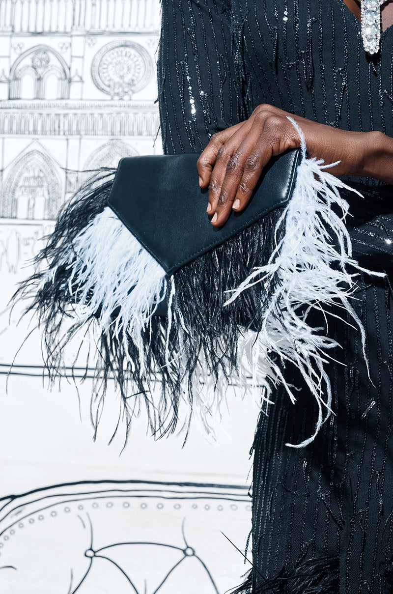 Close-up of a person holding the MIXED DECISIONS FEATHER CLUTCH, composed of faux leather and decorated with black and white feathers. The person is dressed in a black sequined outfit, and the clutch includes a magnetic snap button closure. The background reveals a sketched architectural design.