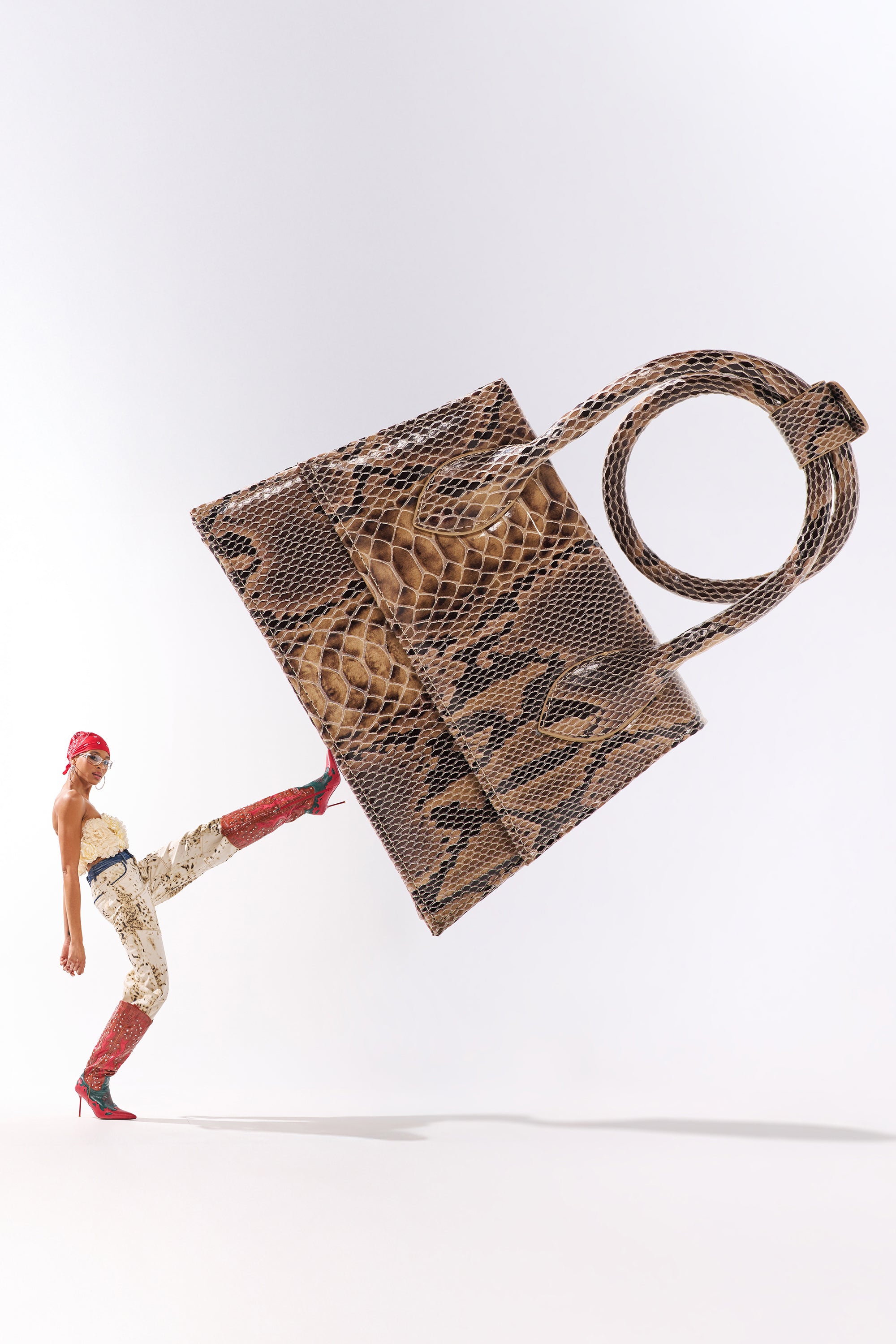 A woman in patterned pants, red boots, and a red headscarf kicks her leg up, showcasing the structured IN THE WILD PURSE with a circular handle and faux leather snakeskin design against a plain white background. 1