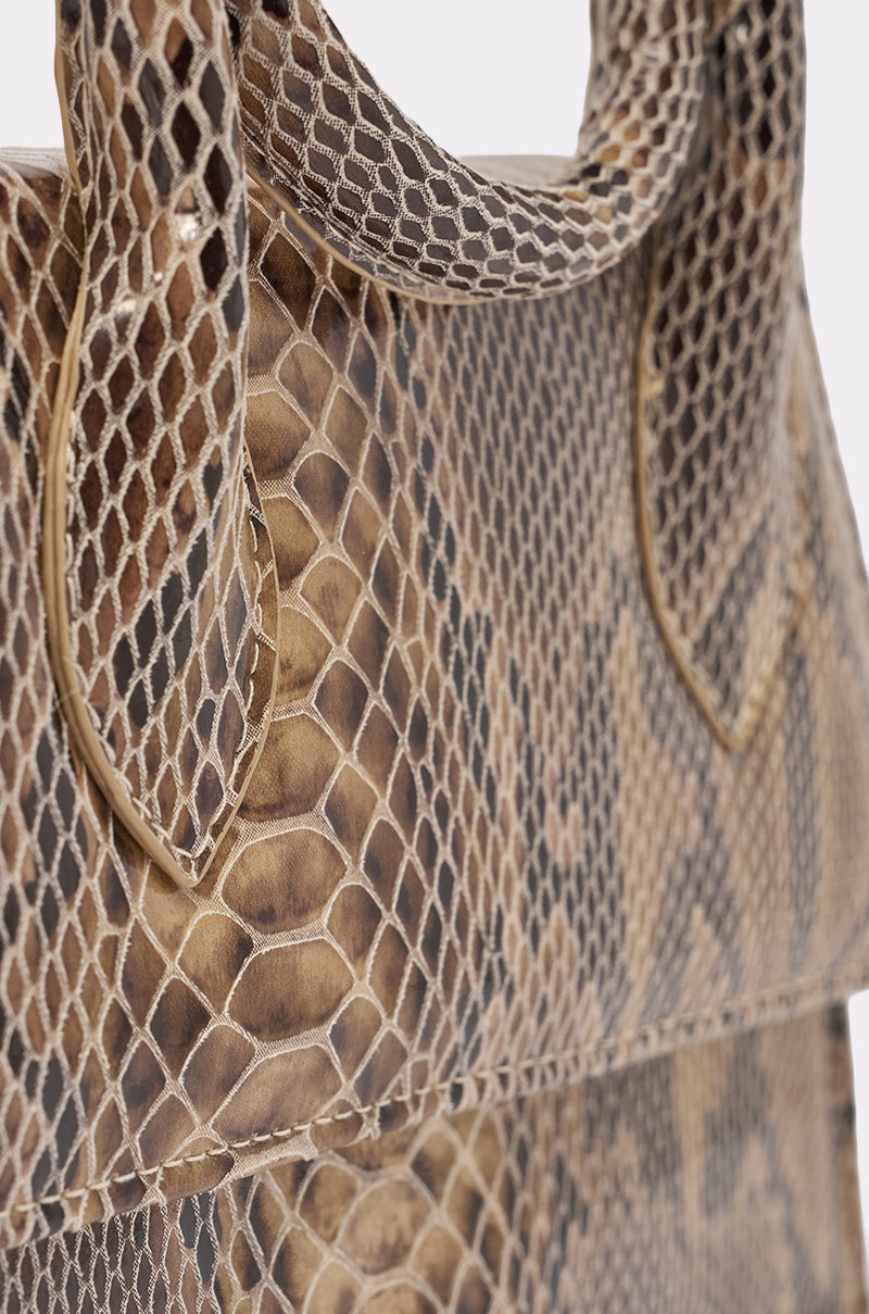 Close-up of the IN THE WILD PURSE, a faux leather snakeskin handbag with a structured shape, detailed scale pattern, and partial view of the handle against a plain background. 6