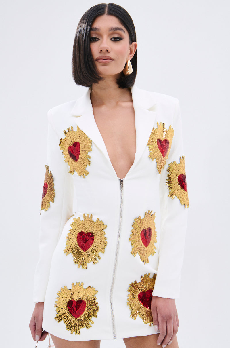 A woman with a sleek bob wears the GLISTENING IN GOLD BLAZER DRESS IN WHITE, featuring a plunging neckline, front zipper, and large red and gold heart-shaped sequin patches, against a plain light background.