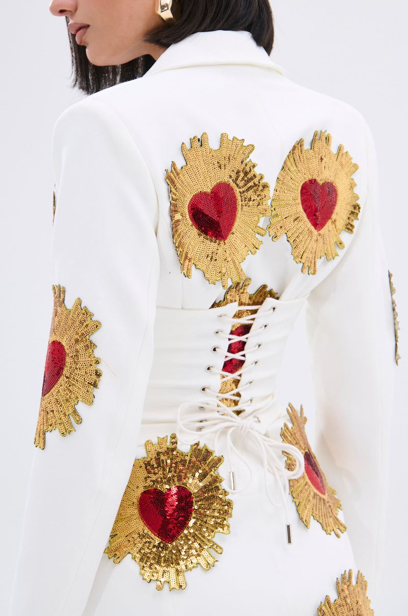 A woman wears the GLISTENING IN GOLD BLAZER DRESS IN WHITE, featuring gold and red sequin patches and a lace-up corset back, seen from behind and slightly to the side.