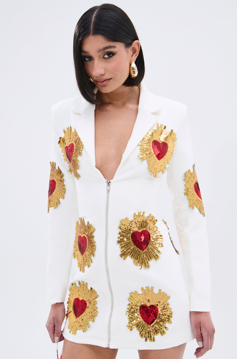 A woman wears the GLISTENING IN GOLD BLAZER DRESS IN WHITE, featuring a plunging neckline, front zipper, and large red and gold sequin patches. She has straight dark hair and accessorizes with gold earrings.