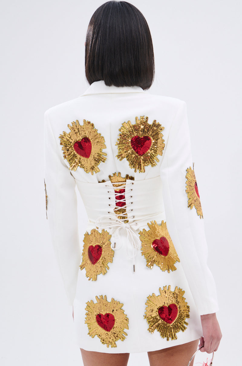 A woman with straight dark hair wears the GLISTENING IN GOLD BLAZER DRESS IN WHITE, featuring a plunging neckline, gold and red sequin heart patches, a lace-up corset back with gold hearts. The backdrop is plain white.
