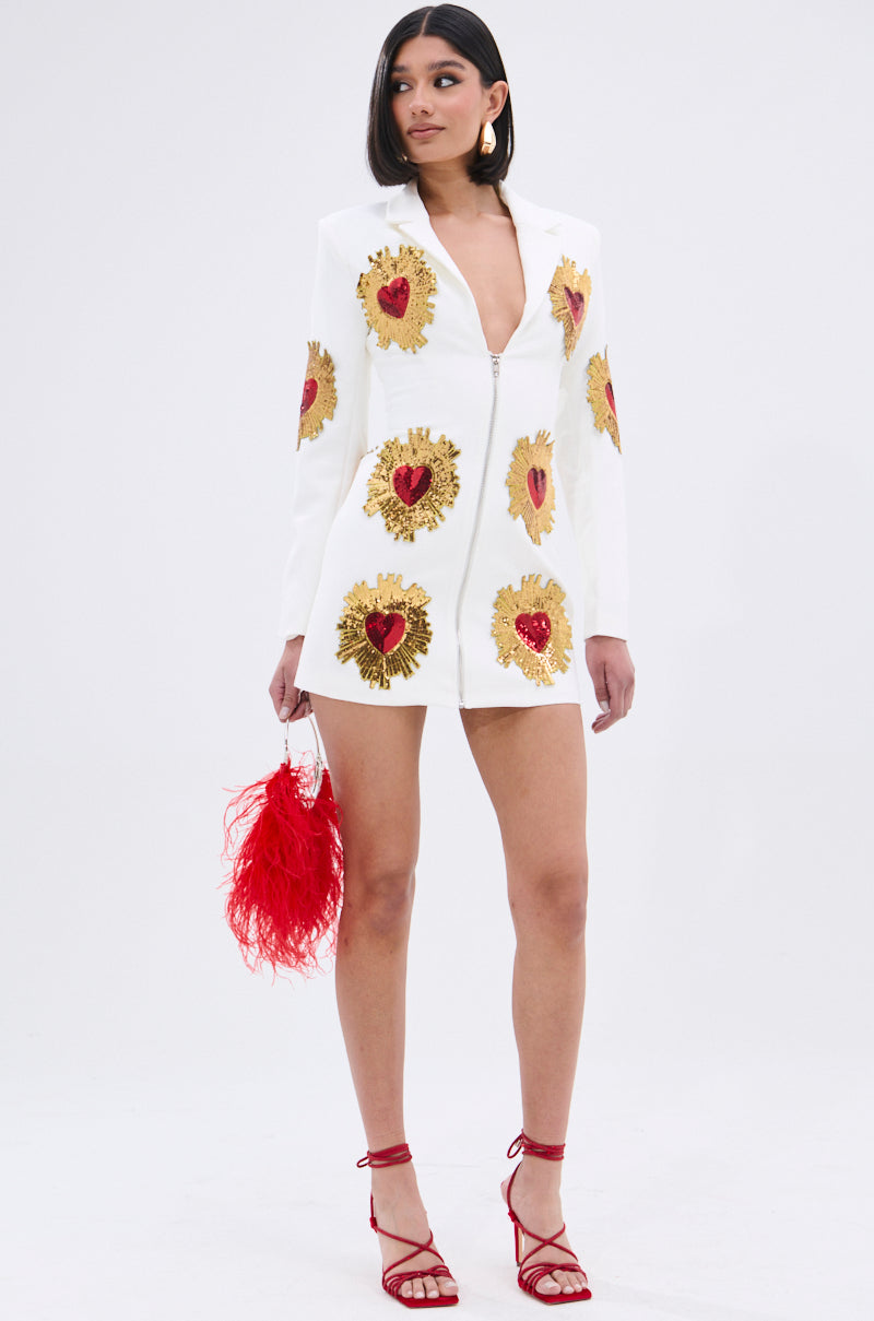 A woman stands against a plain white background wearing the GLISTENING IN GOLD BLAZER DRESS IN WHITE, featuring a plunging neckline and heart-shaped sequin patches, paired with a red feathery handbag and red high-heeled sandals.