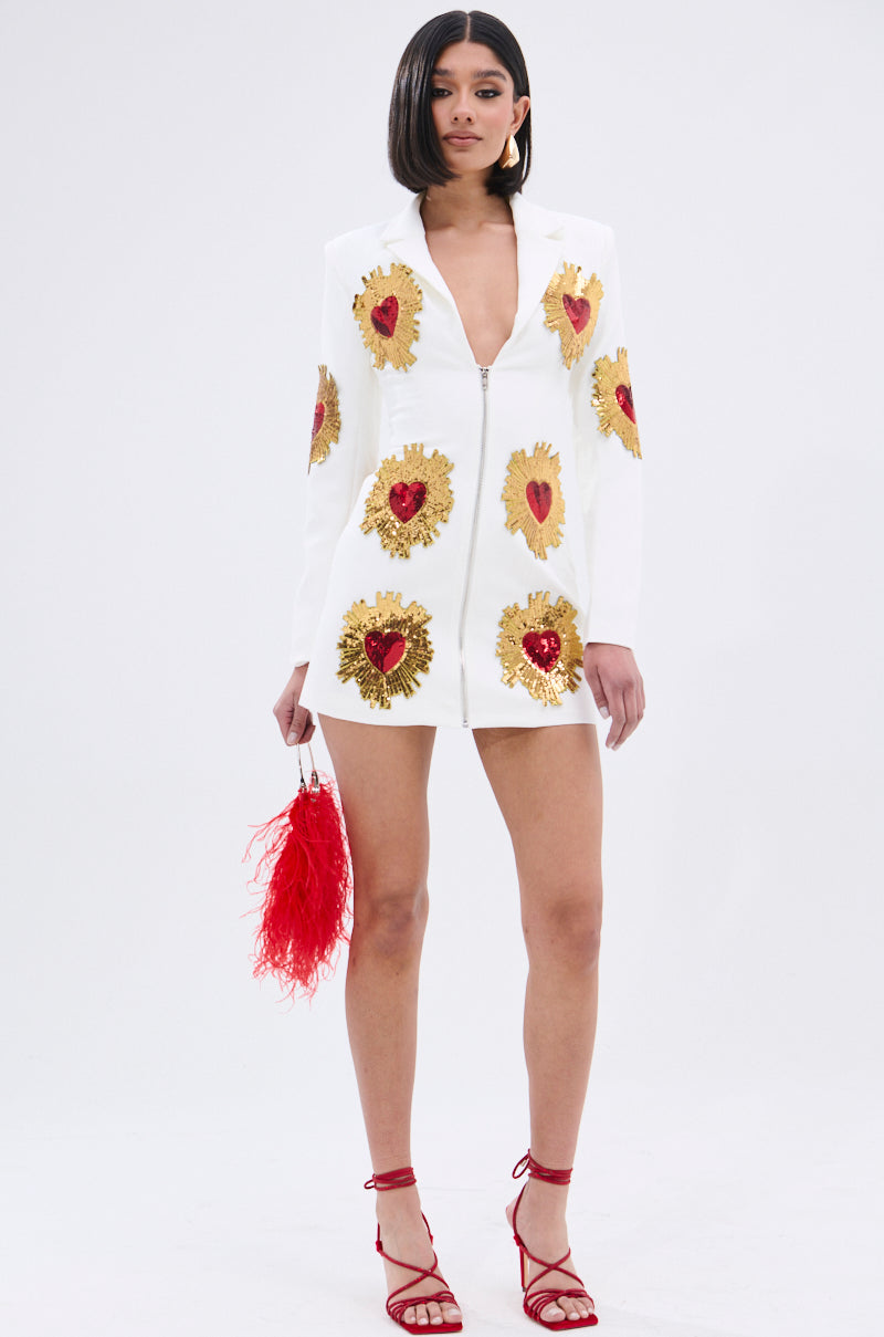 A woman wearing the GLISTENING IN GOLD BLAZER DRESS IN WHITE with gold and red heart-shaped sequin patches, red strappy heels, and a red feathered handbag stands against a plain background.