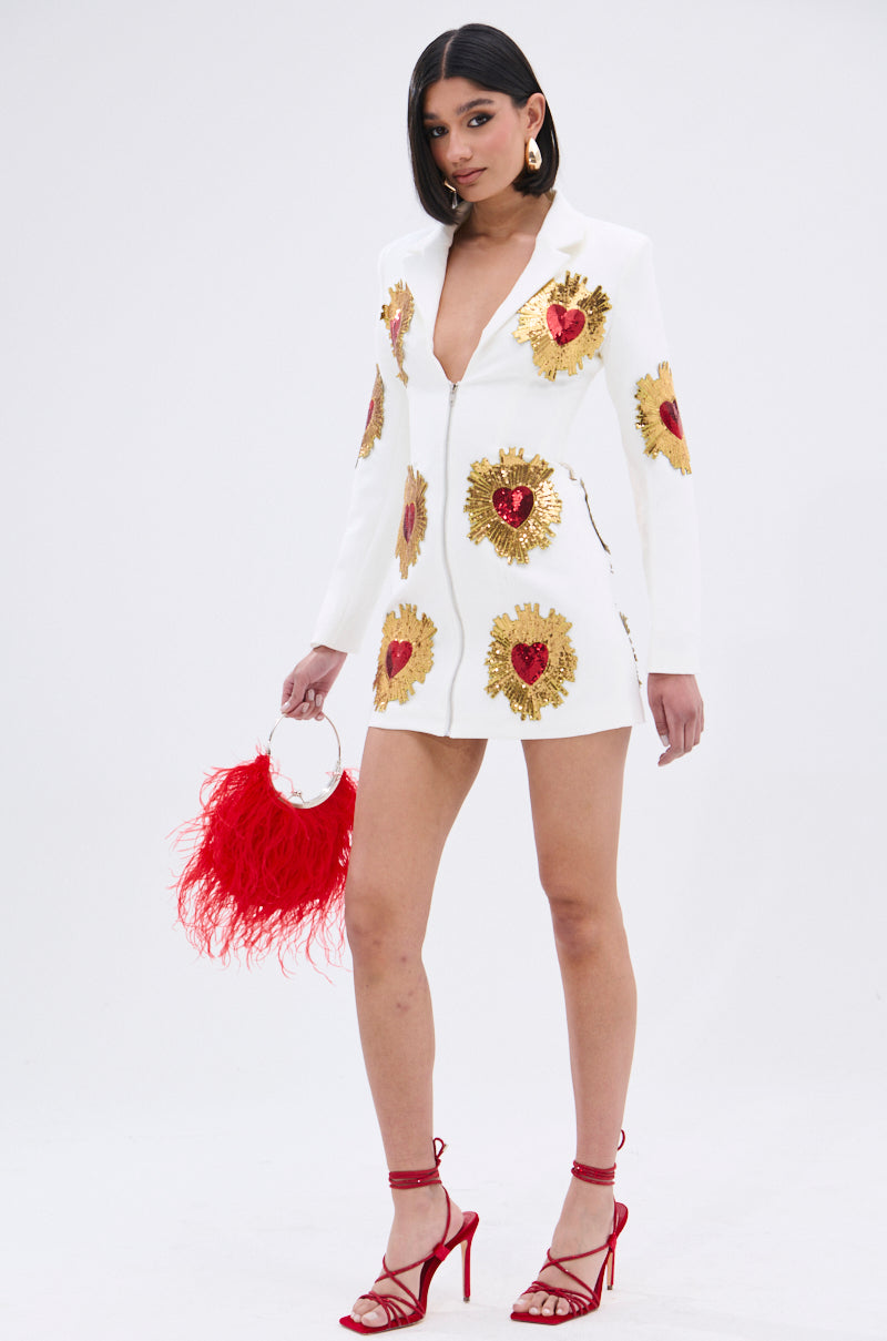 A woman poses in the GLISTENING IN GOLD BLAZER DRESS IN WHITE with a plunging neckline and gold and red heart sequin patches, holding a red feathered handbag and wearing red high-heeled sandals against a plain background.