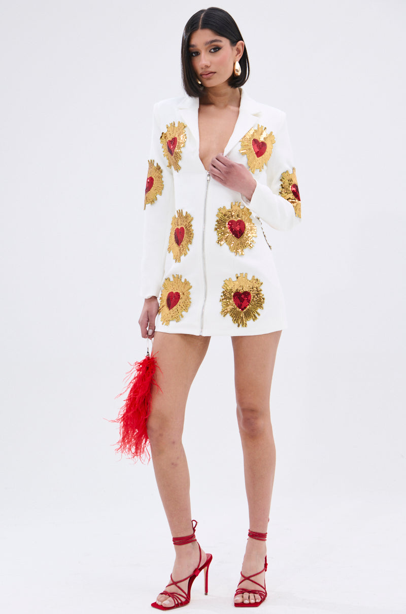 A woman in a GLISTENING IN GOLD BLAZER DRESS IN WHITE, featuring a plunging neckline and sequin patches with red heart designs, stands against a plain background holding a red feathered clutch and wearing red strappy high heels.