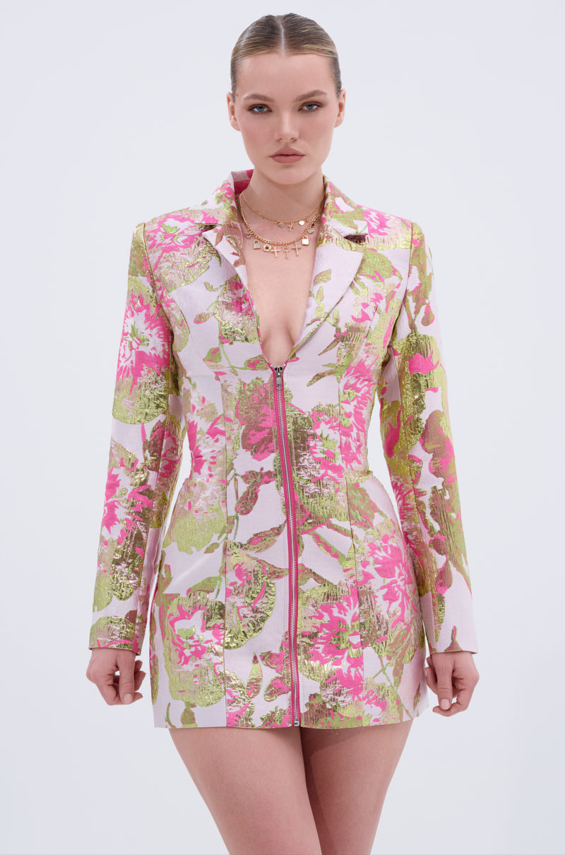 A woman wears the PRETTY IN PINK BROCADE BLAZER DRESS, a fitted, long-sleeved dress with a pink, green, and gold floral pattern, deep V-neckline, and front zipper. She looks forward with a neutral expression.