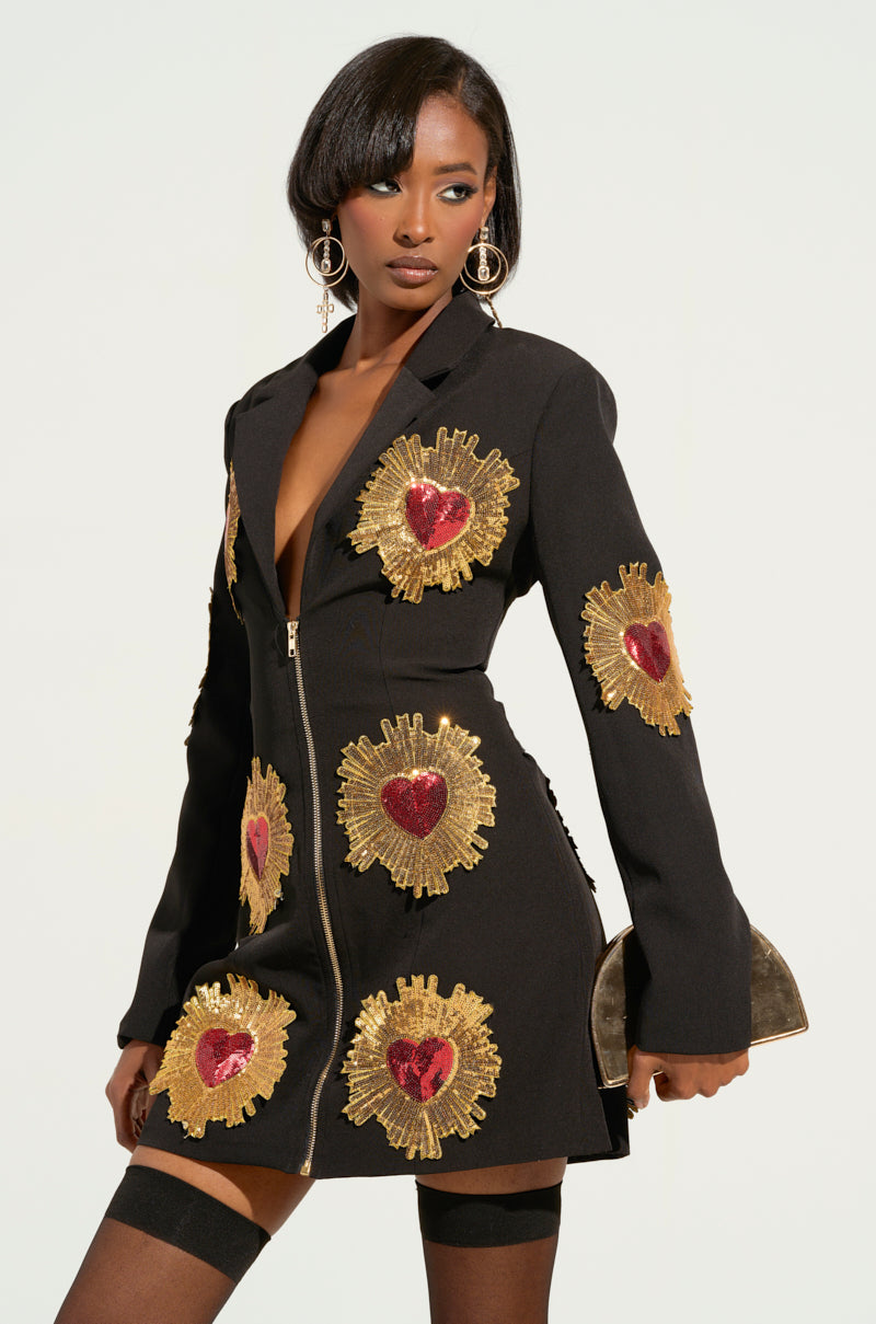 A person wearing the GLISTENIN IN GOLD BLAZER DRESS, adorned with sequin sunburst detailing, stands against a light background. They accessorize with a metallic clutch, large hoop earrings, thigh-high stockings, and heart-shaped accents to complete the look.