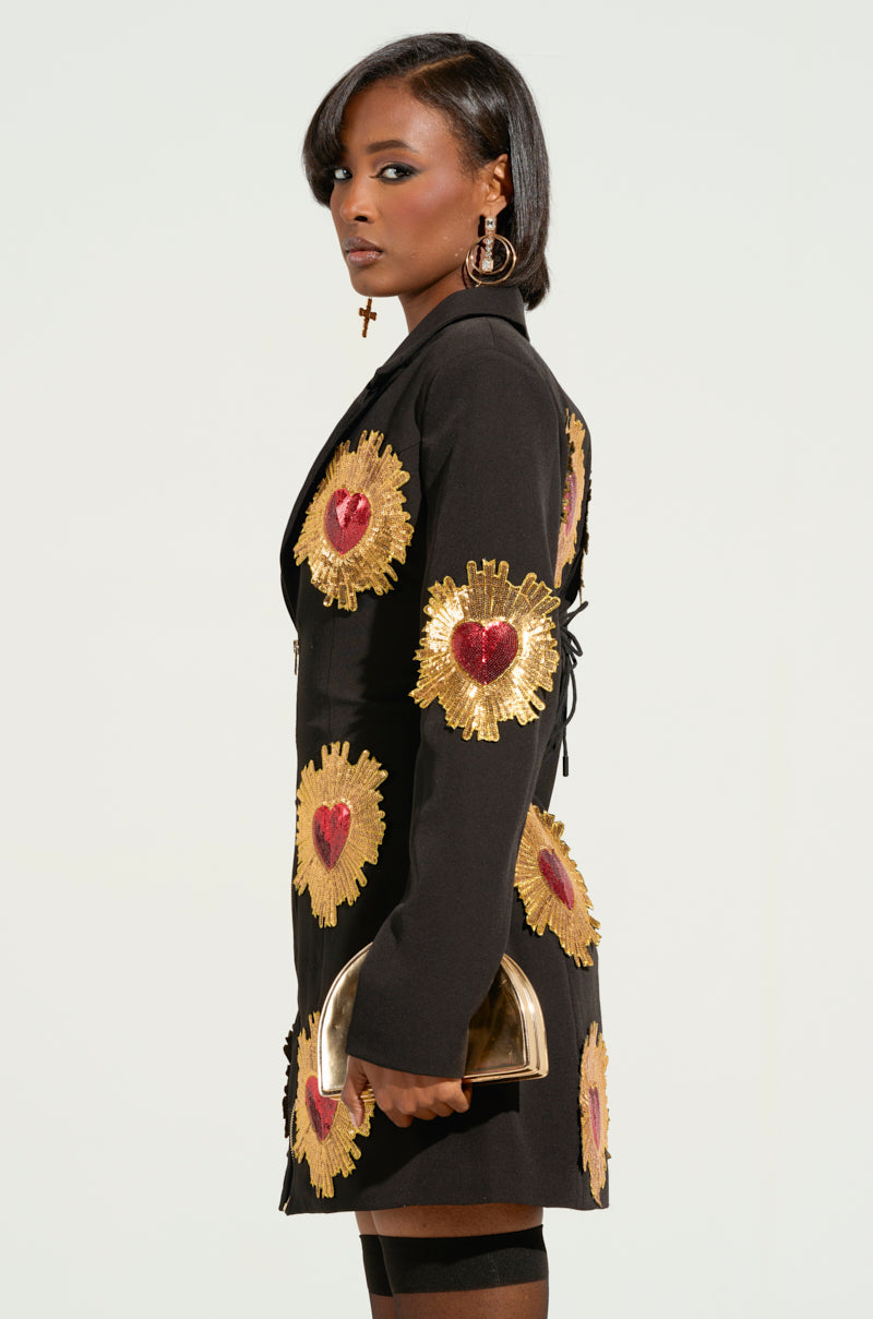 Wearing the GLISTENIN IN GOLD BLAZER DRESS, a person stands confidently in a stylish ensemble characterized by its black base and gold and red heart-shaped patterns. Complementing this look is a shiny metallic handbag and statement jewelry featuring large hoop earrings, a cross-shaped earring, all highlighted by elegant, sequin sunburst detailing.