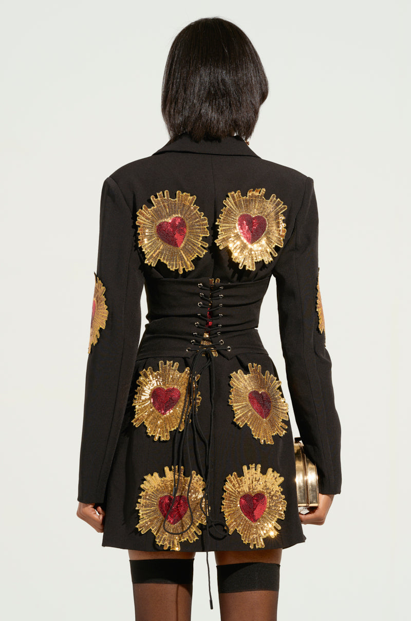 With their back to the camera, an individual wears the GLISTENIN IN GOLD BLAZER DRESS, featuring gold and red heart embellishments against a black backdrop. The dress is elegantly designed with a lace-up back panel that cinches the waist. They complement this captivating look by holding a small round clutch.