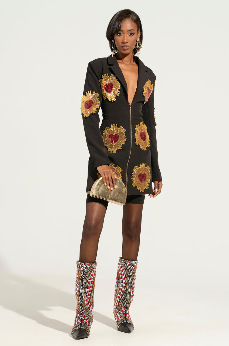 Dressed in the GLISTENIN IN GOLD BLAZER DRESS adorned with golden sunflower and red heart designs, a woman poses gracefully. She complements her ensemble with a small gold clutch and patterned knee-high boots, while hoop earrings add an extra touch of elegance to her look against the plain white background that enhances her chic style.