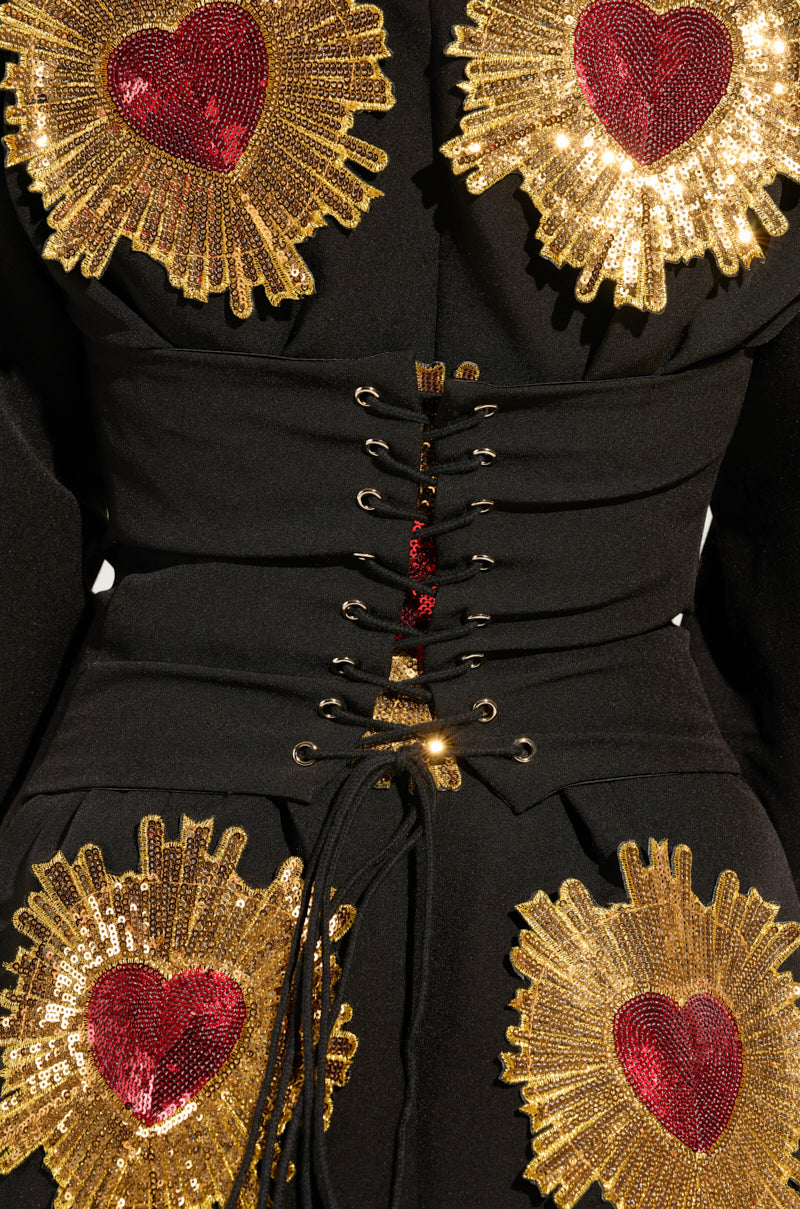A close-up of a black corset dress highlights sequin sunburst detailing centered with red hearts, alongside a lace-up front design featuring gold eyelets and black laces, beautifully enhancing the style of the GLISTENIN IN GOLD BLAZER DRESS.