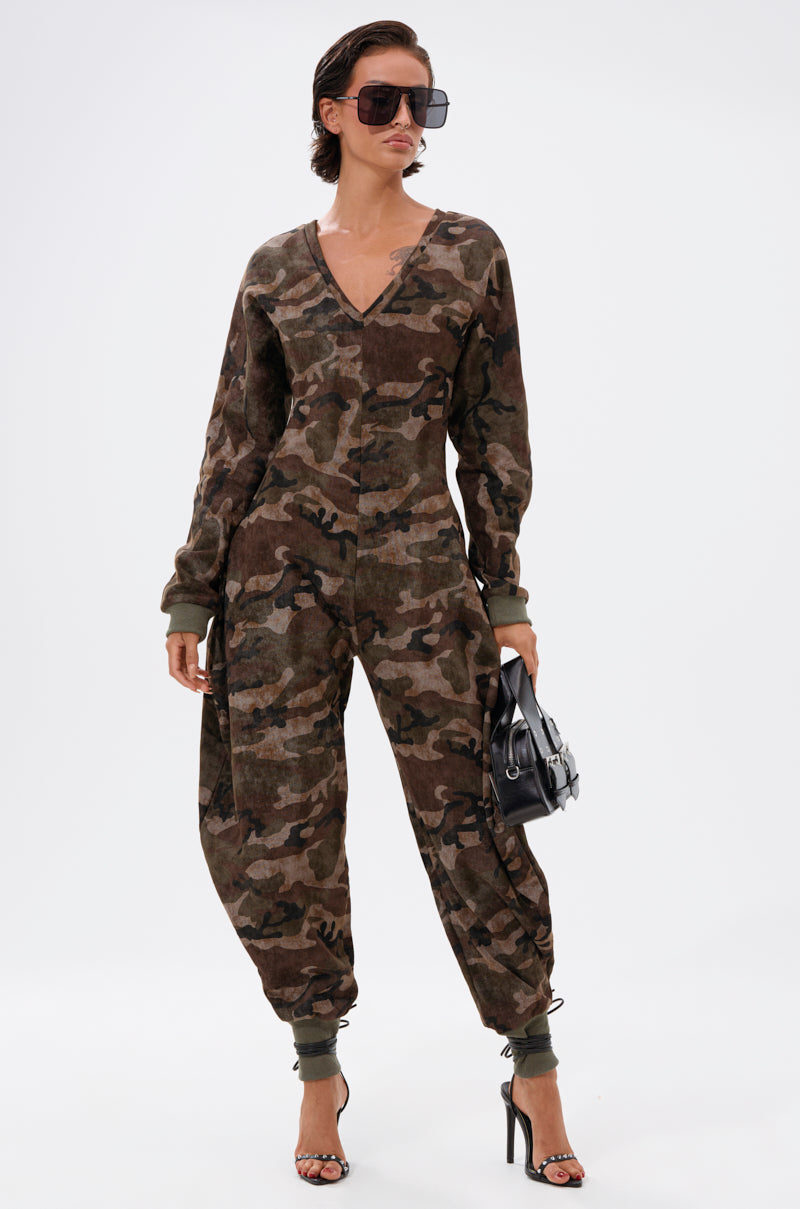 A woman stands confidently against a white background, wearing the COMFY COZY SWEATSHIRT JUMPSUIT IN CAMO, black sunglasses, high-heeled sandals, and holding a small black handbag.