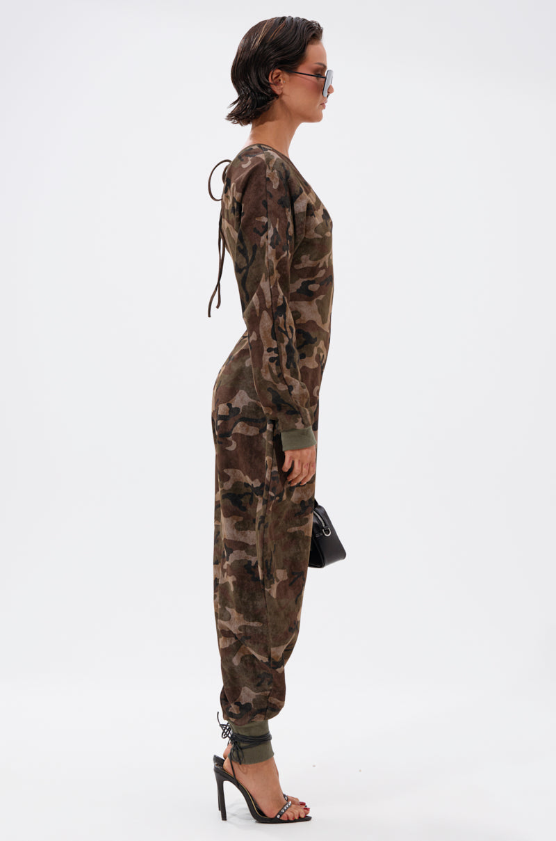 Woman in profile stands against a white background, wearing the COMFY COZY SWEATSHIRT JUMPSUIT IN CAMO with long sleeves, paired with black high-heeled sandals, sunglasses, and a small black handbag.